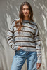 A woman wearing a gray and brown horizontally striped knit sweater with light blue jeans, standing against a textured beige wall with leaf shadows.