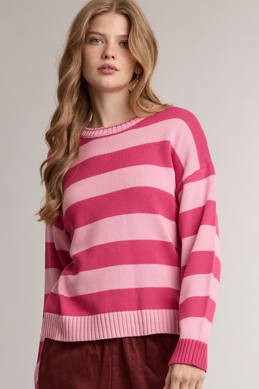 A woman wearing a pink and light pink horizontally striped knit sweater, paired with burgundy pants, against a neutral light gray studio background.