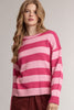 A woman wearing a pink and light pink horizontally striped knit sweater, paired with burgundy pants, against a neutral light gray studio background.