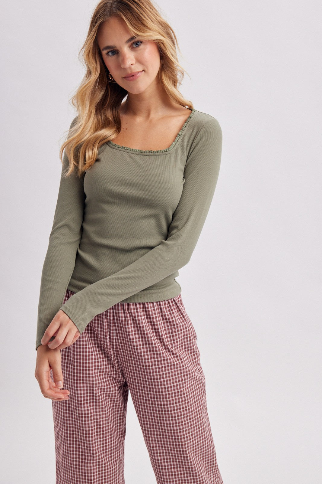 A woman modeling a long-sleeve olive green top with a scoop neckline and red-and-white gingham check pants against a light gray studio background.