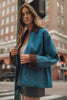 A woman wearing a blue denim jacket with brown faux-leather trim stands on a city street corner.