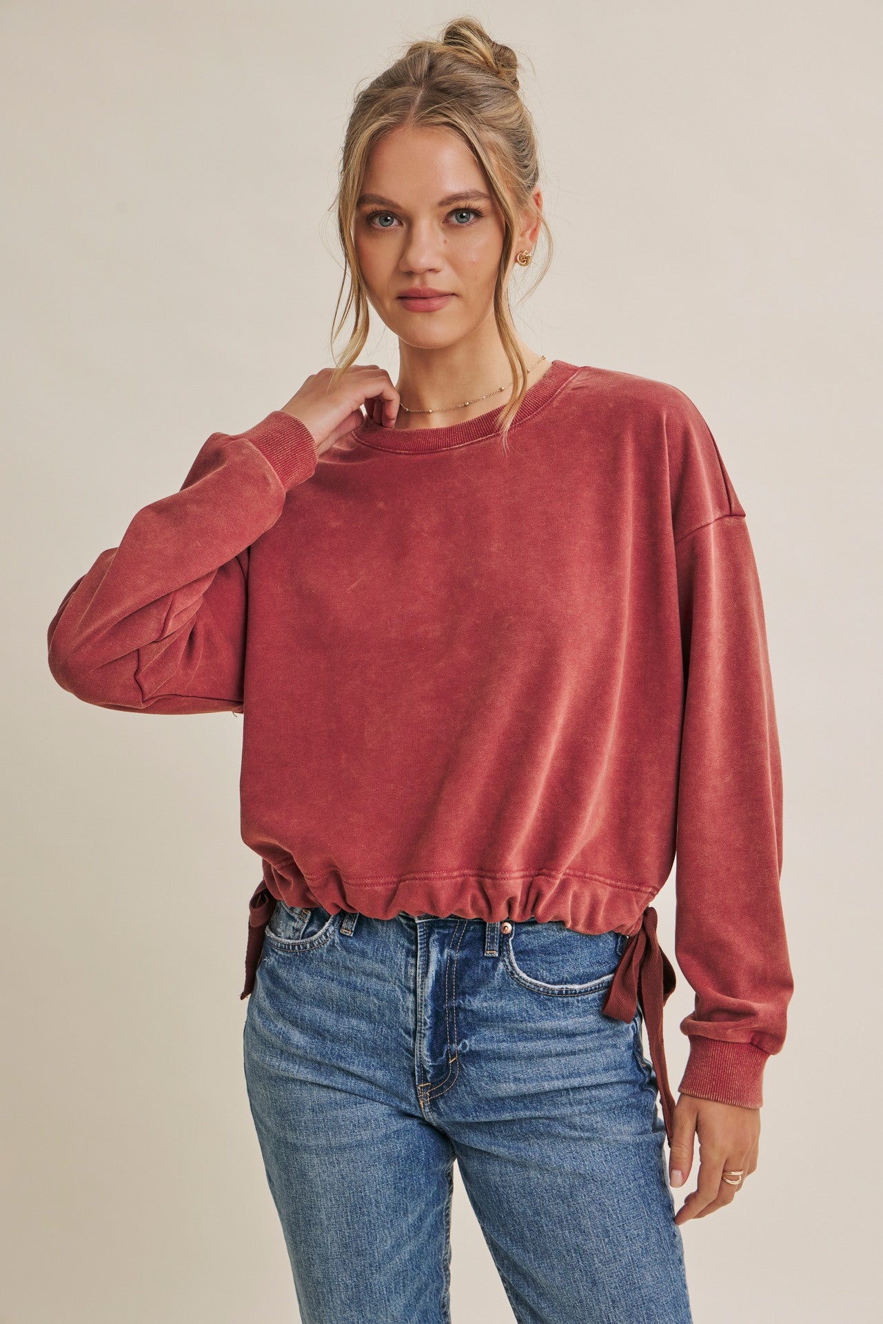 A woman wearing a dusty rose velvet long-sleeve sweatshirt with side ties and blue jeans, posing against a solid beige studio background.