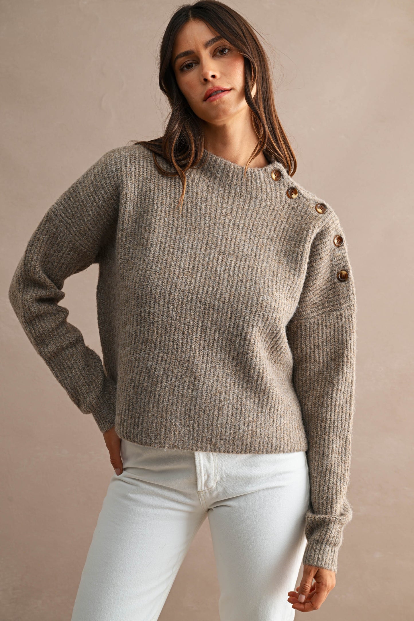 A woman wearing a ribbed knit sweater with decorative brown buttons on the shoulders, paired with white pants, against a plain beige studio background.