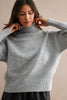 A woman wearing a light gray ribbed knit turtleneck sweater with wide sleeves, posing against a neutral beige background.
