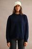 A woman wearing a dark navy blue fuzzy knit crewneck sweater, dark gray jeans, and a light cream baseball cap, standing against a textured beige studio background.