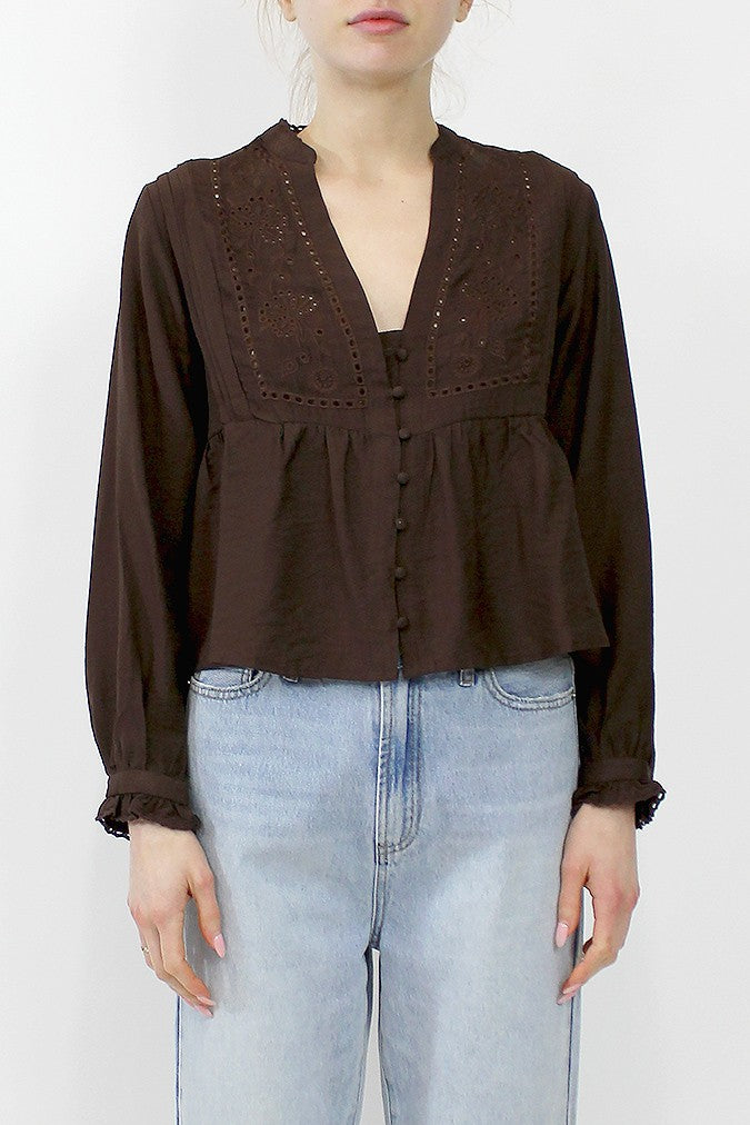 A woman wearing a dark brown long-sleeve blouse with a lace-up front and eyelet embroidery on the chest, paired with light-wash blue jeans, against a plain white background.
