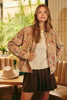 A woman stands in a cafe setting wearing a beige floral button-up jacket over a cream ribbed sweater and a dark brown pleated skirt. A light beige hat rests on a wooden table in the foreground.