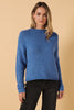 A woman wearing a chunky blue knit sweater and black jeans, standing against a solid beige background.