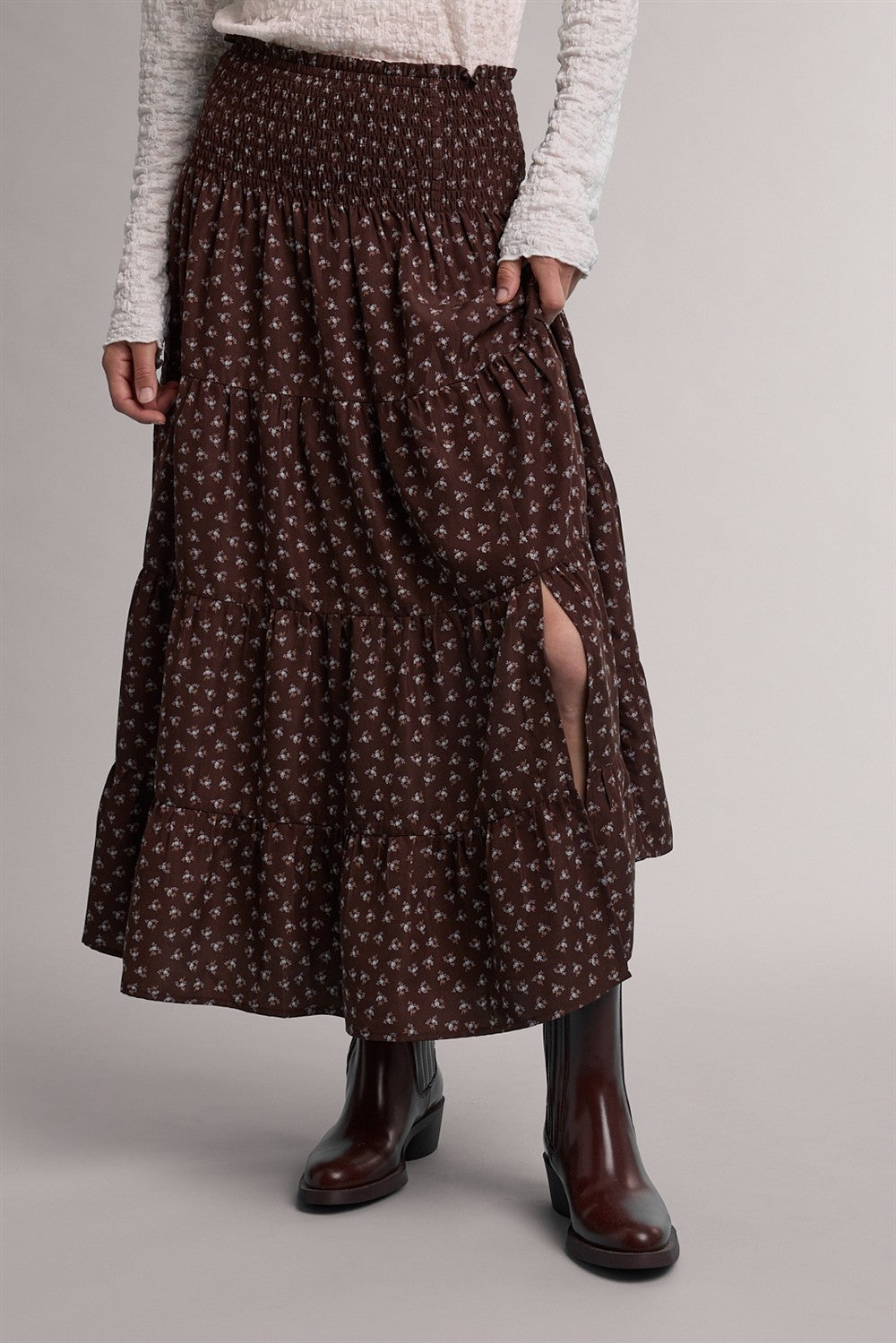 A dark brown smocked midi skirt with a small white floral pattern, featuring a side slit and pockets, styled with a cream knit top and dark brown ankle boots.