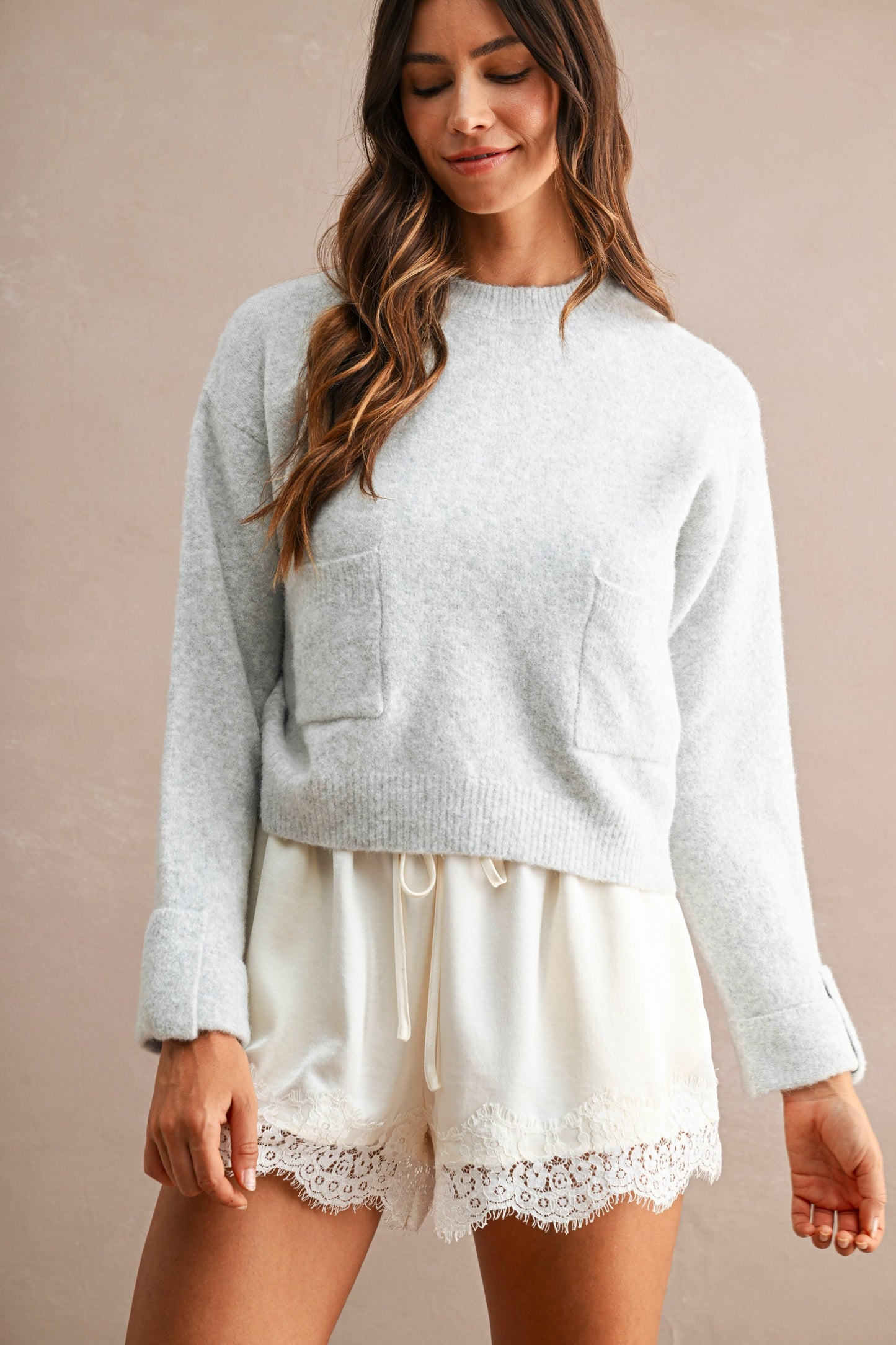 A woman wearing a light gray knit crewneck sweater with front patch pockets and cream-colored shorts with lace trim, standing against a solid beige background.