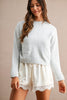 A woman wearing a light gray knit crewneck sweater with front patch pockets and cream-colored shorts with lace trim, standing against a solid beige background.