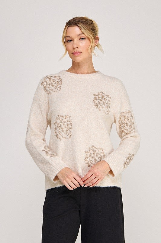 A woman modeling a cream-colored knit sweater with beige floral patterns on the chest and sleeves, paired with black pants, against a light gray studio background.