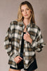 A woman modeling an oversized long-sleeve plaid button-up shirt in brown, beige, and white, worn open over a black top and dark denim shorts against a neutral studio backdrop.