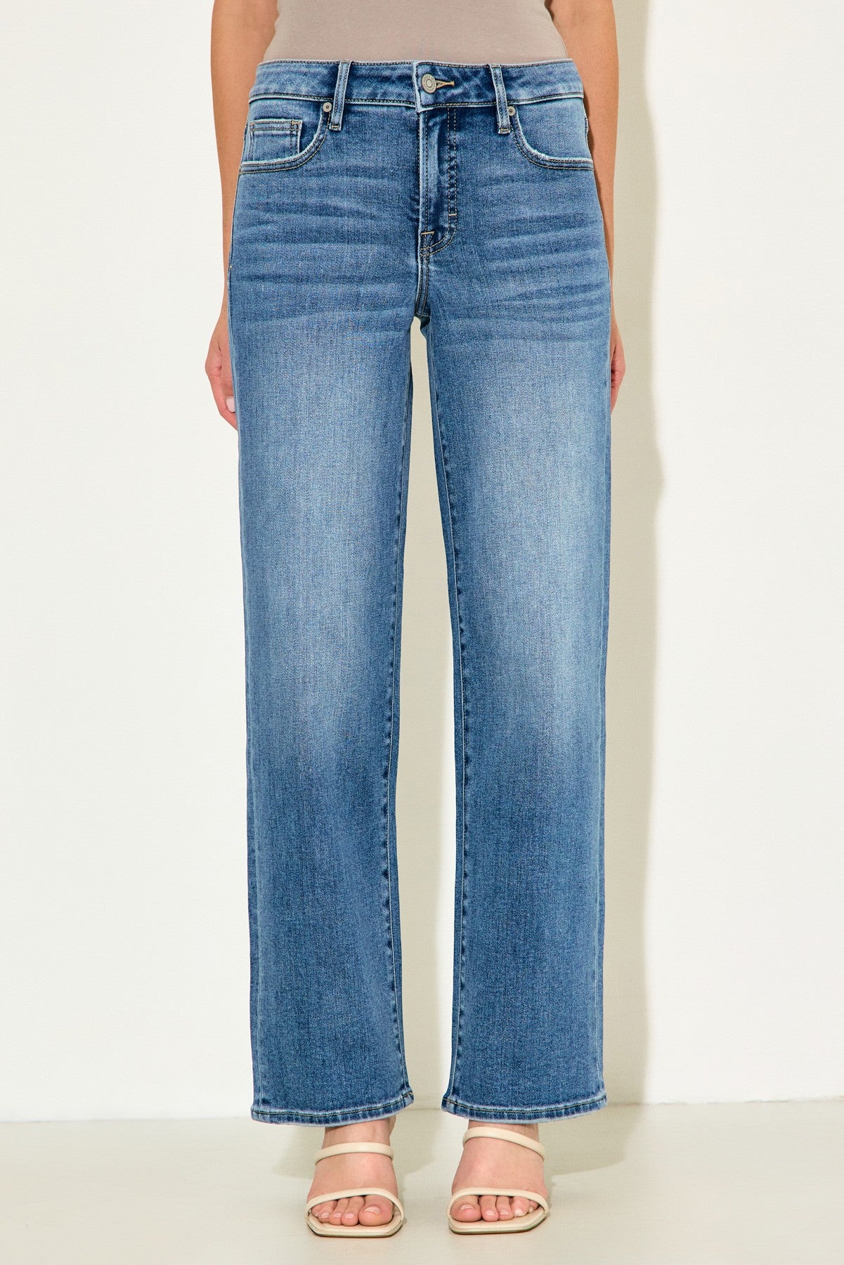 A model wearing a pair of high-waisted, wide-leg blue denim jeans with a faded wash, paired with beige heeled sandals against a plain off-white background.