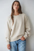 A woman wearing a cream-colored knit crewneck sweater and light blue jeans, standing against a plain light gray wall.