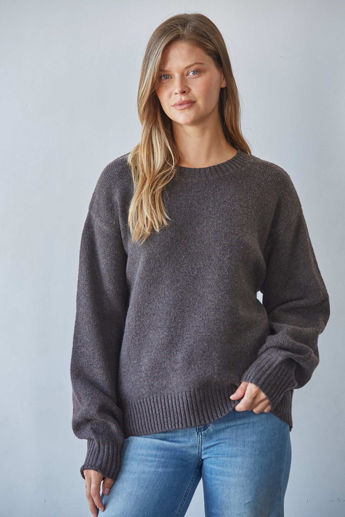 A woman wearing a dark gray crewneck knit sweater and blue jeans, standing against a light gray studio background.