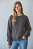 A woman wearing a dark gray crewneck knit sweater and blue jeans, standing against a light gray studio background.
