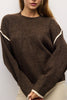 A woman wearing a brown knit sweater with contrasting cream-colored trim along the shoulders and cuffs, paired with light beige pants.