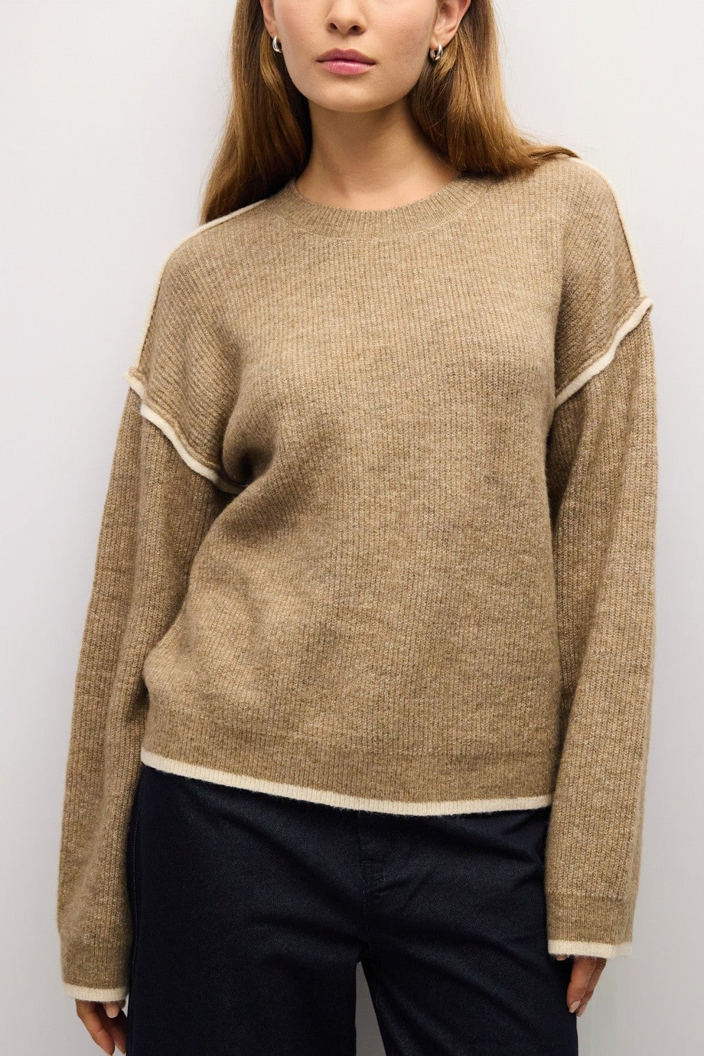A woman wearing a beige ribbed knit sweater with contrasting cream trim along the shoulders, hem, and cuffs, paired with dark pants against a light gray background.