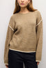 A woman wearing a beige ribbed knit sweater with contrasting cream trim along the shoulders, hem, and cuffs, paired with dark pants against a light gray background.