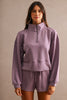 A woman modeling a matching lavender half-zip sweatshirt and shorts set against a neutral textured beige background.