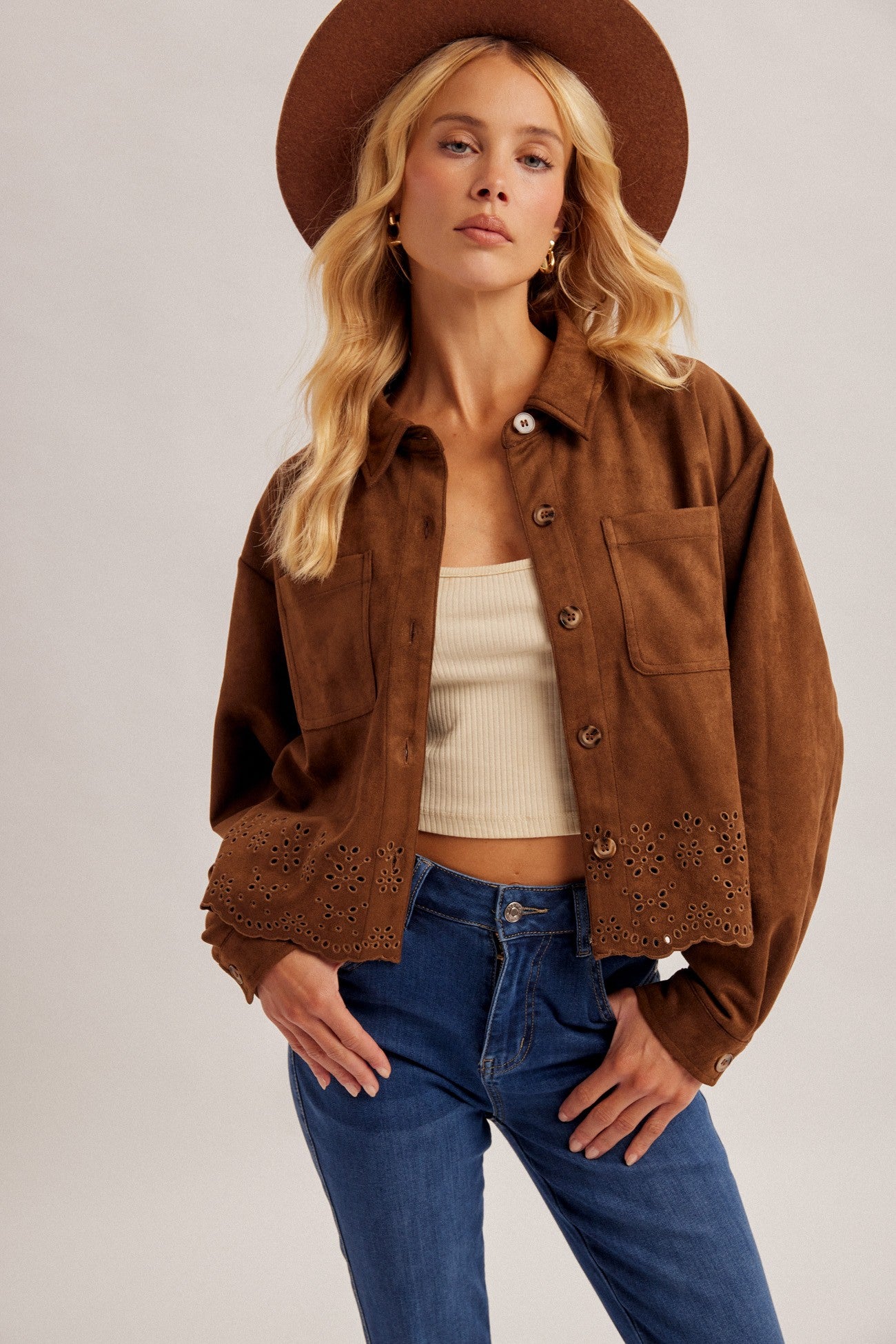 A woman modeling a cropped brown suede-like button-up shirt jacket with eyelet lace detailing at the cuffs, paired with blue jeans, a cream ribbed tank top, and a brown wide-brim hat against a light gray studio background.