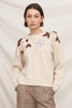 A person wearing a cream knit sweater with brown and beige floral embroidery and a light blue flower motif on the chest, paired with brown plaid trousers, against a soft off-white fabric backdrop.