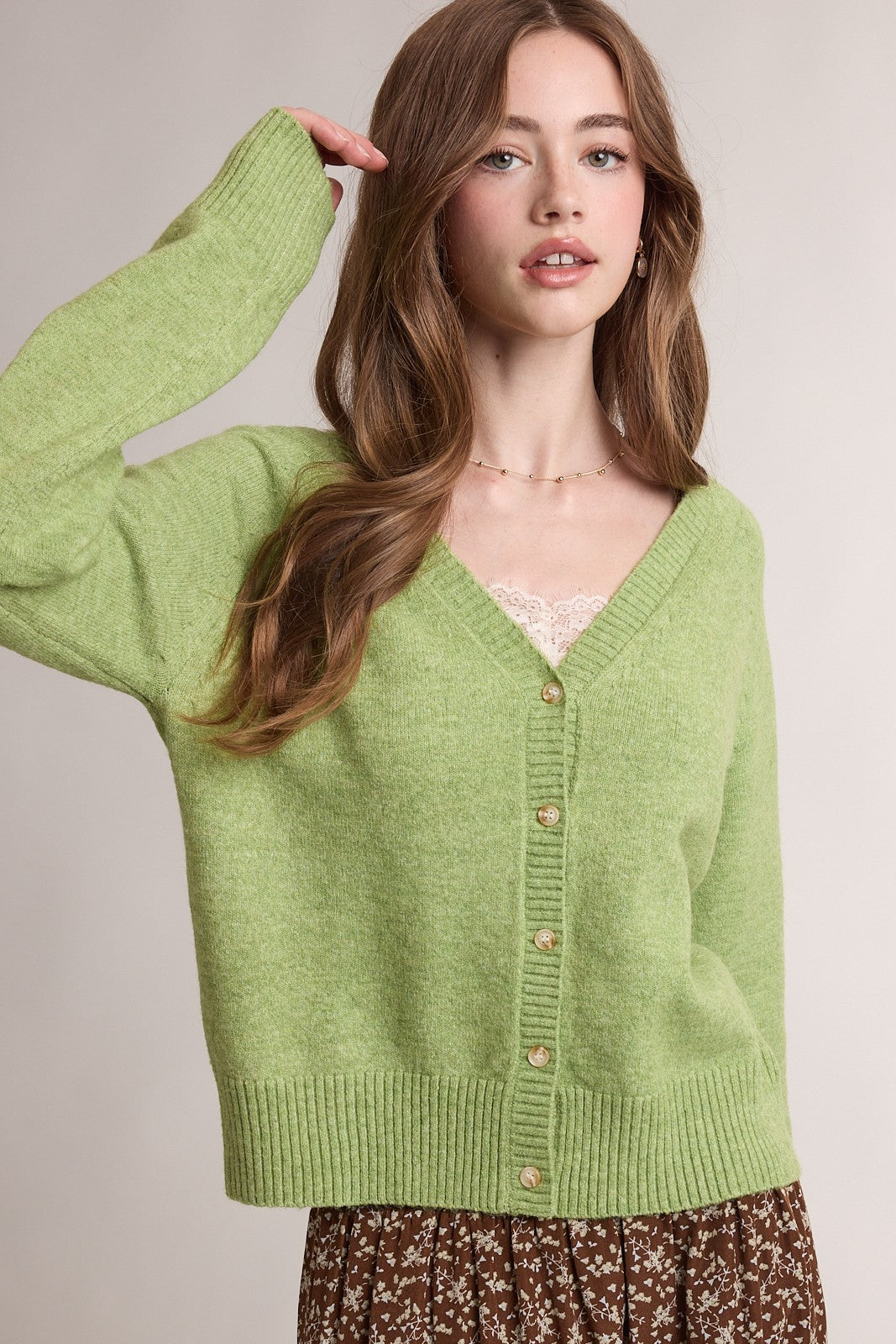 A woman wearing a light green knit cardigan sweater with brown buttons, styled with a lace top and a brown floral skirt, against a neutral beige background.