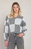 A woman wearing a gray and white geometric checkered knit sweater paired with gray jeans, standing against a solid light beige background.