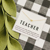 Gold necklace with a heart pendant on a card labeled 'TEACHER' on a checkered background.