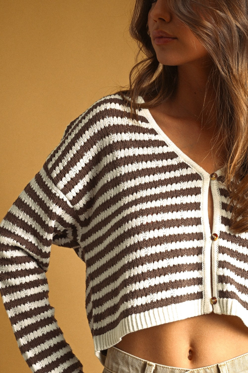 A woman wearing a brown and cream striped cropped cardigan sweater, shown from the side against a solid warm tan background.