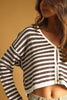 A woman wearing a brown and cream striped cropped cardigan sweater, shown from the side against a solid warm tan background.