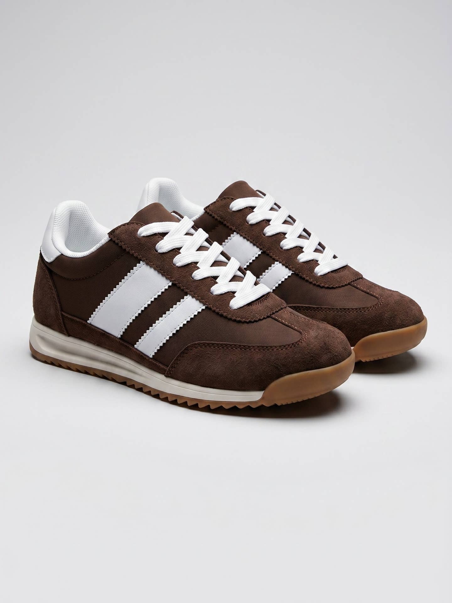A pair of brown suede sneakers with three white stripes on the side, white laces, and a gum sole, photographed on a light gray studio background.