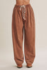 A person wearing loose, wide-leg brown corduroy pants with an elastic drawstring waist, shown from the waist down against a plain light beige studio background.