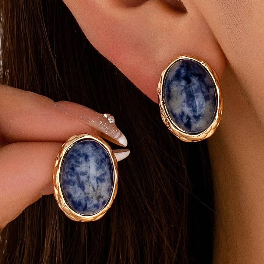 Close-up of a person wearing and holding a pair of oval stud earrings with blue marbled stones in textured gold settings, with dark brown hair and skin visible.