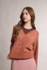 A woman modeling a rust-orange knit cardigan sweater with a brown V-neck undershirt and a pink polka-dot skirt, photographed against a neutral light gray studio background.