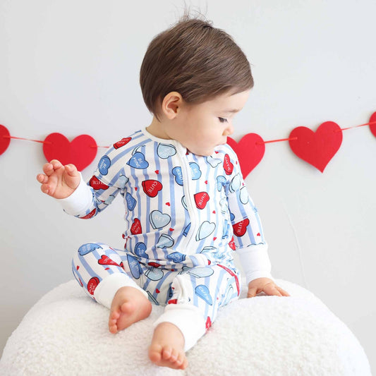 Child wearing a heart-patterned onesie sitting on a white cushion with red heart decorations in the background.
