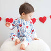 Child wearing a heart-patterned onesie sitting on a white cushion with red heart decorations in the background.