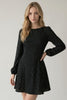A woman modeling a black long-sleeve fit-and-flare dress with a small white polka dot pattern against a light gray studio background.