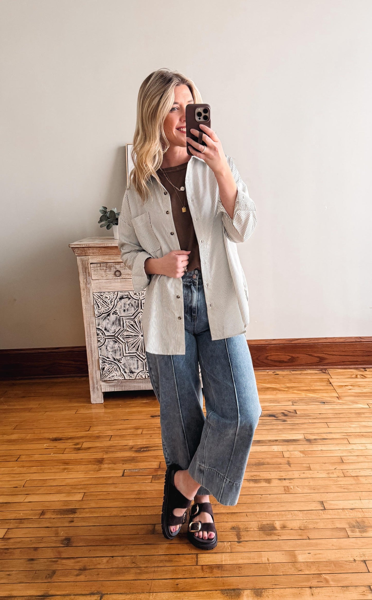Woman taking a mirror selfie wearing a light-colored jacket and blue jeans in a room with wooden flooring.