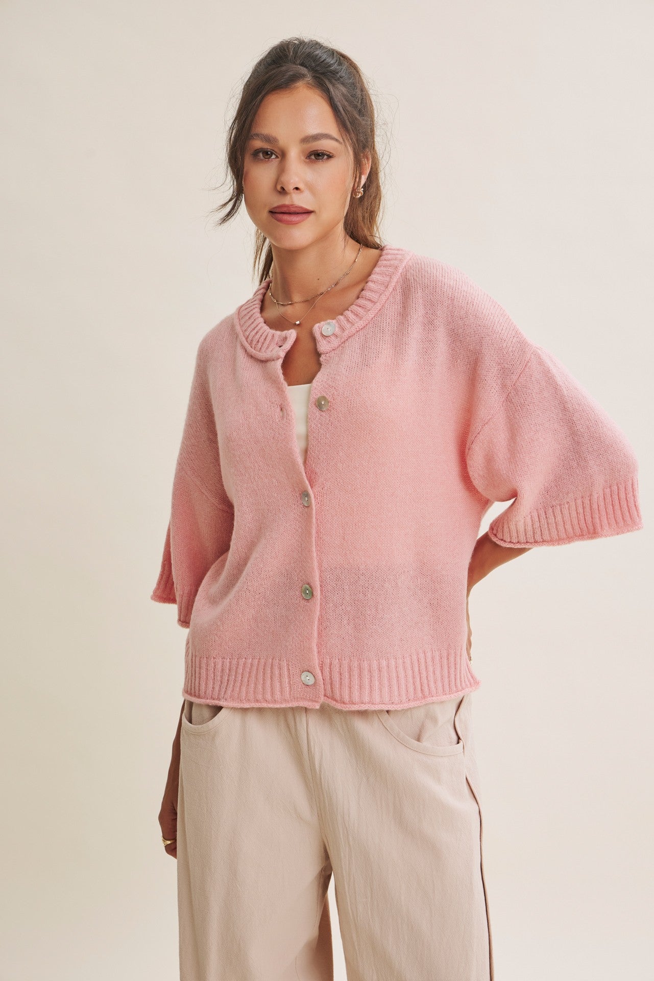 A woman wearing a light pink knit cardigan with short dolman sleeves, a round collar, and front buttons, paired with light beige pants, against a plain beige studio background.