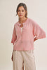 A woman wearing a light pink knit cardigan with short dolman sleeves, a round collar, and front buttons, paired with light beige pants, against a plain beige studio background.