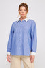 A woman modeling a blue and white vertically striped button-down shirt with a solid white collar and cuffs, paired with beige pants, against a light gray studio background.