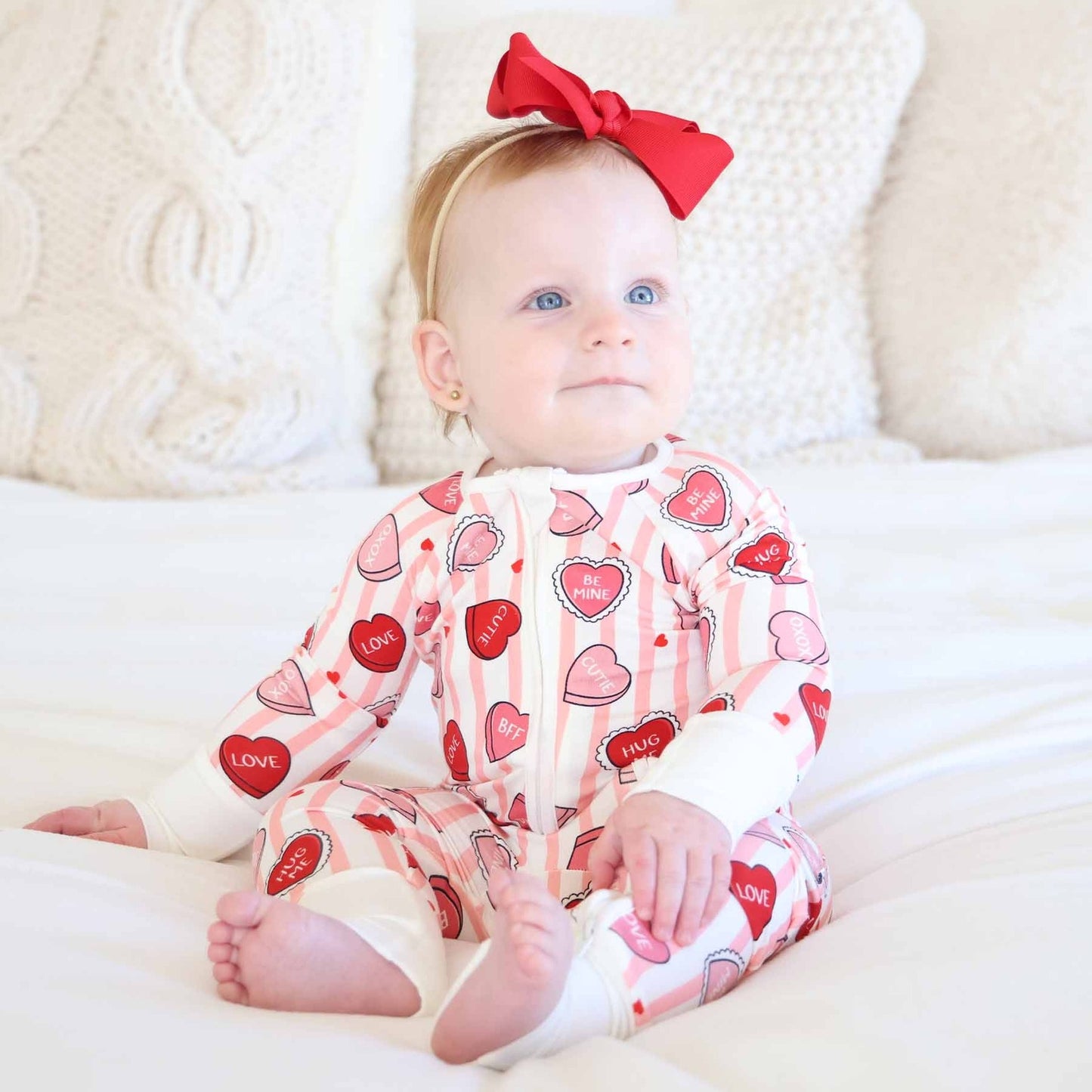 Baby wearing a Valentine's Day-themed onesie with hearts and 'LOVE' text, sitting on a white surface.