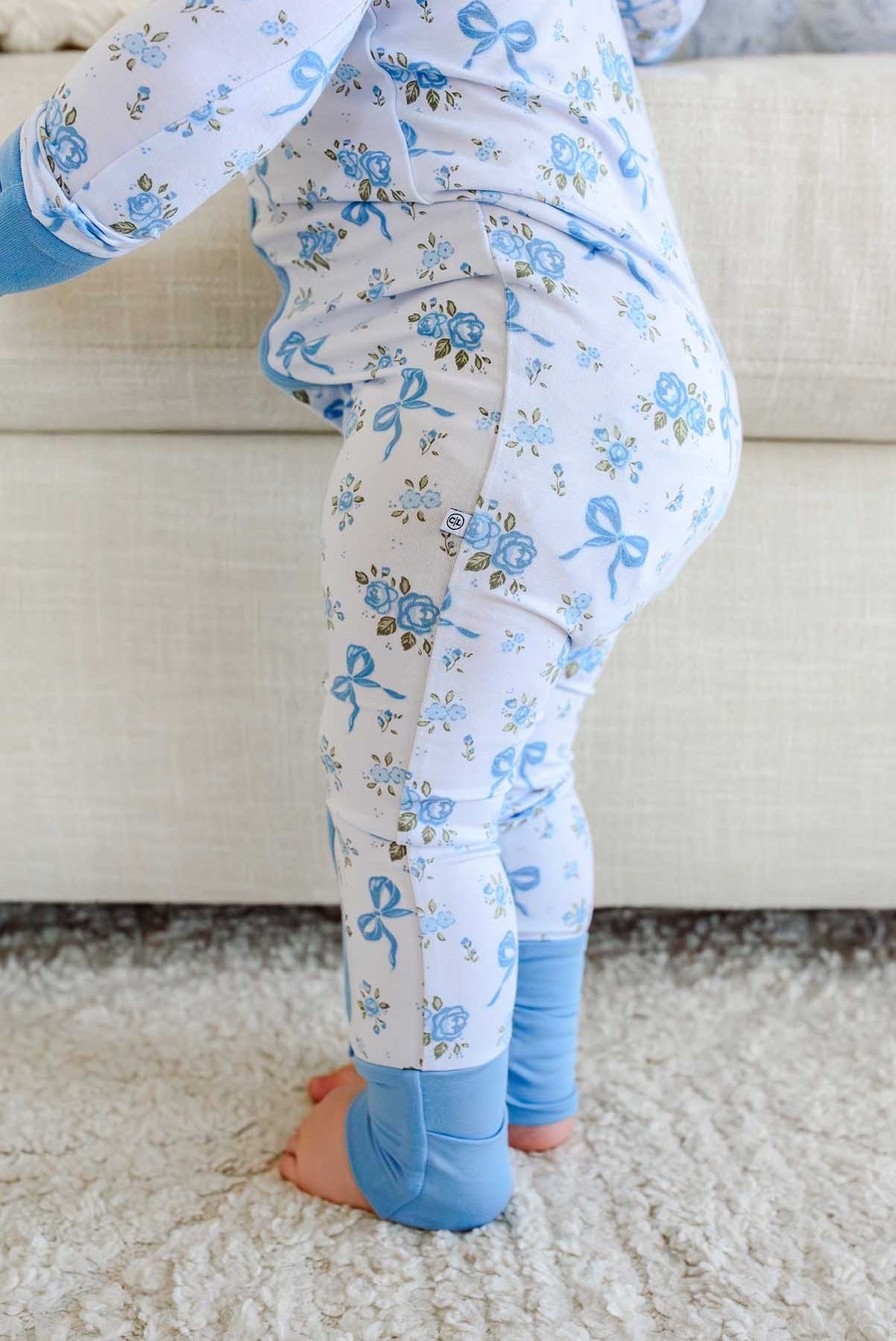 A close-up of a baby wearing light blue and white floral pajama pants with blue leg warmers, standing on a white shag rug in front of a beige sofa.