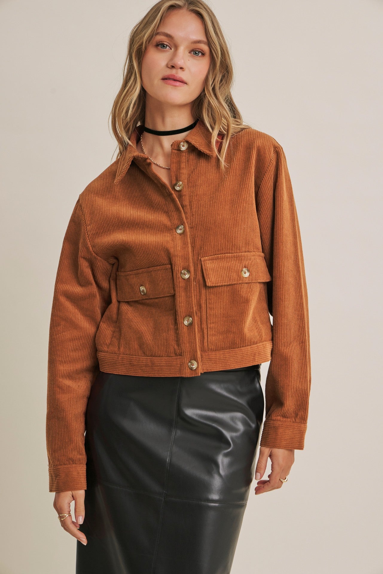 A woman wearing a cropped brown corduroy jacket with button details and flap pockets, paired with a black faux-leather skirt, against a plain beige background.