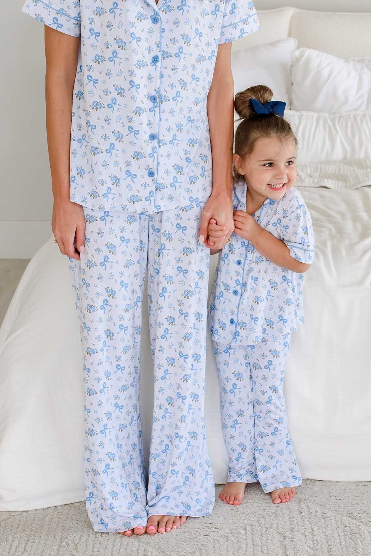 A matching adult and child sleepwear set in a bedroom. The set consists of light blue and white floral pajamas, with the adult standing on a light carpet holding the child's hand.