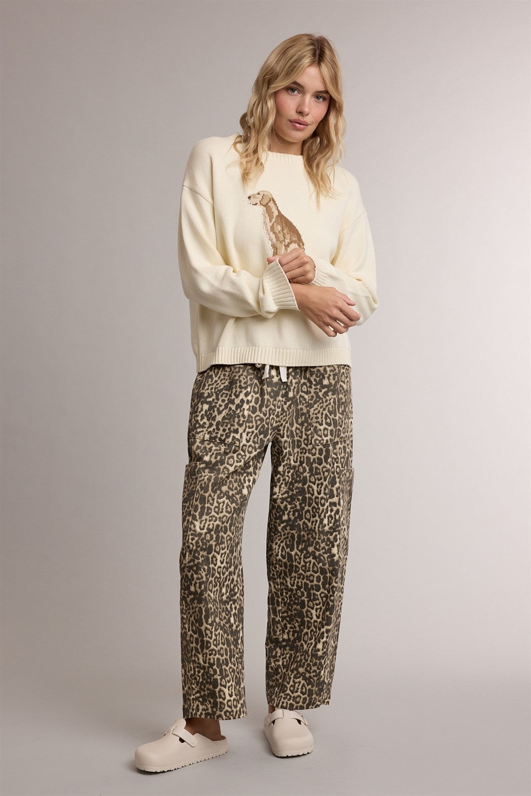 A woman modeling a cream-colored knit sweater with a giraffe graphic and matching leopard-print pants, standing against a neutral light gray studio background.