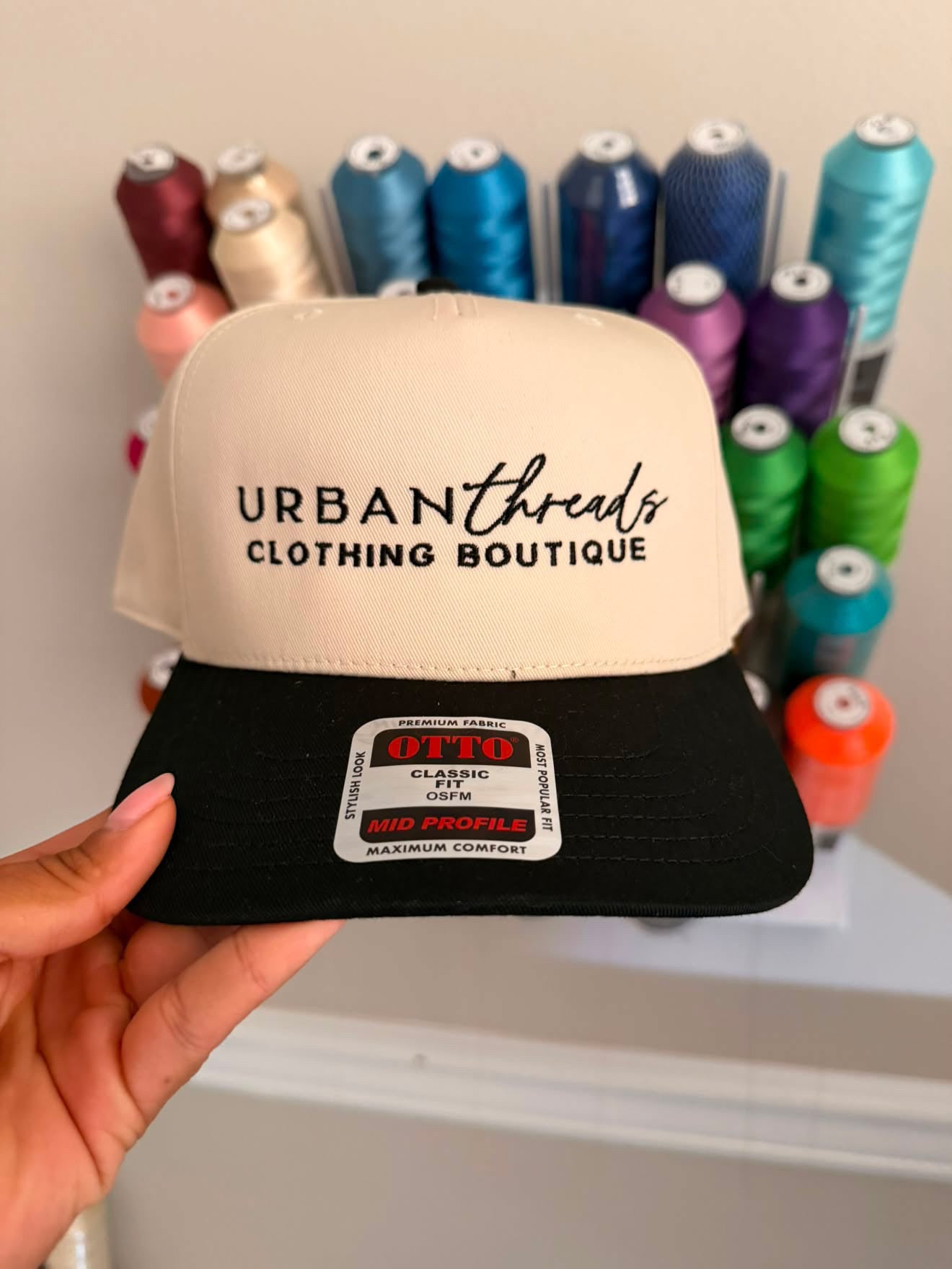 A hand holds a beige and black snapback cap in front of a display of colorful spools of thread. The cap features the text 'URBAN threads CLOTHING BOUTIQUE' on the front panel and a 'OTTO Classic Cap' sticker on the black brim.