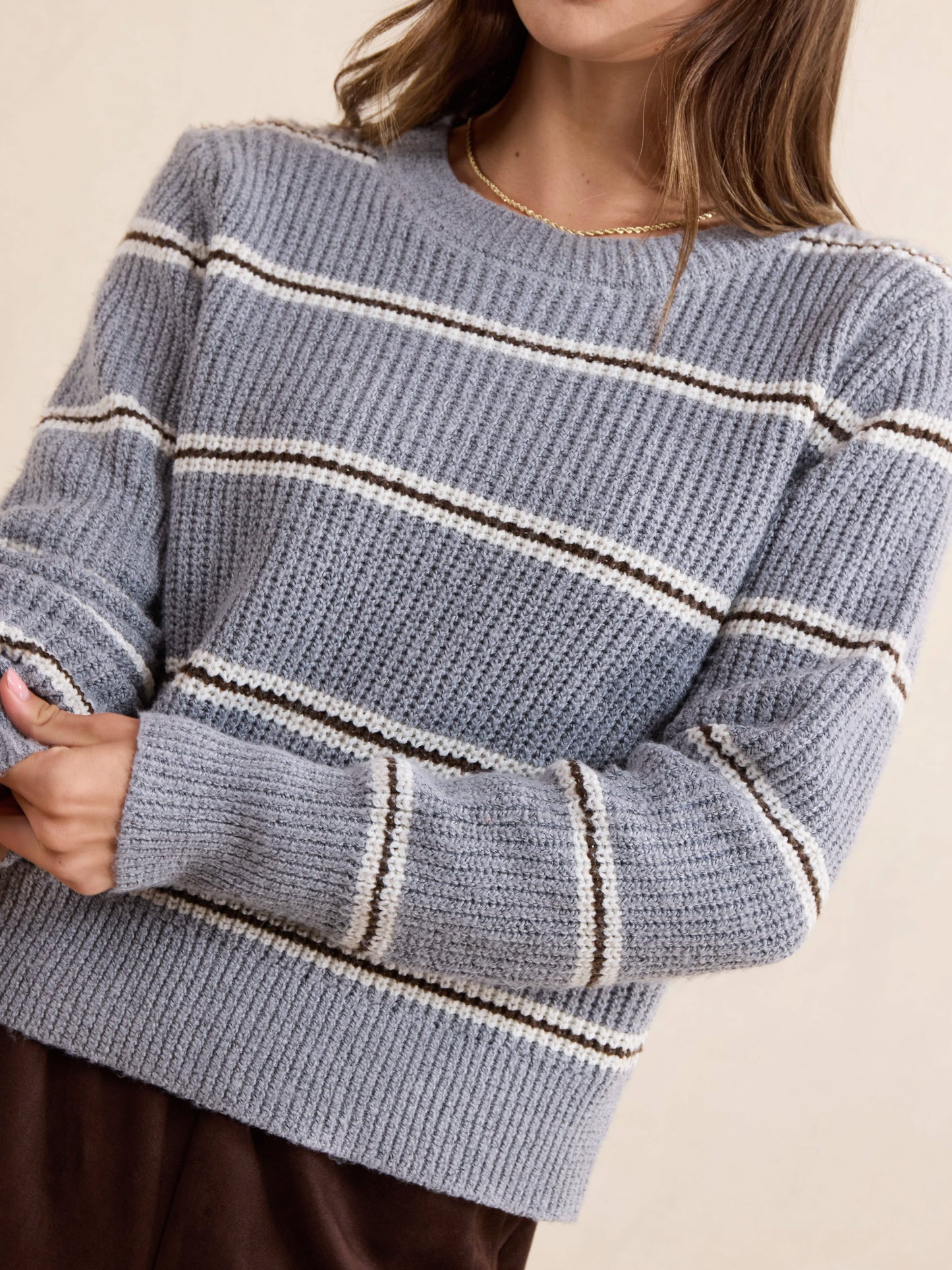 A close-up of a woman wearing a chunky knit crewneck sweater with horizontal stripes in light blue, cream, and dark brown, paired with a gold necklace and brown bottoms.